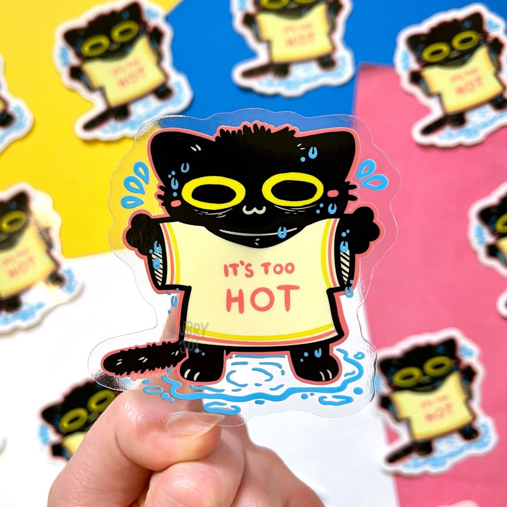 A hand holding a sticker of a black cat wearing an oversized cream colored T-shirt that says “it’s too hot” on it. The cat is T-posing and looks sweaty & frazzled, but is smiling. It’s sweating to a little puddle underneath it. The sticker has clear edges.