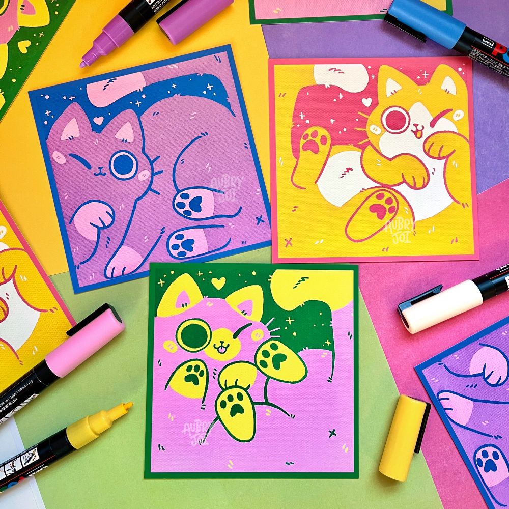 Three art prints of canvas paintings of cute  cats squeezed into the canvas, so that barely any of the backgrounds are showing. One is purple with a blue background/frame; one is yellow with a coral background/frame; the last is pink with a green background/frame. 