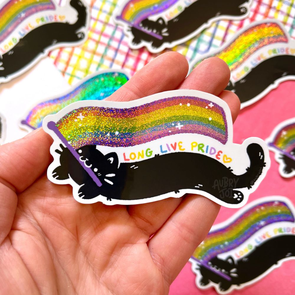A hand holding a sticker of a wiggly long black cat holding a glittery, wiggly long rainbow pride flag in its mouth. Over the cat’s back, it says “long live Pride” in rainbow colors.