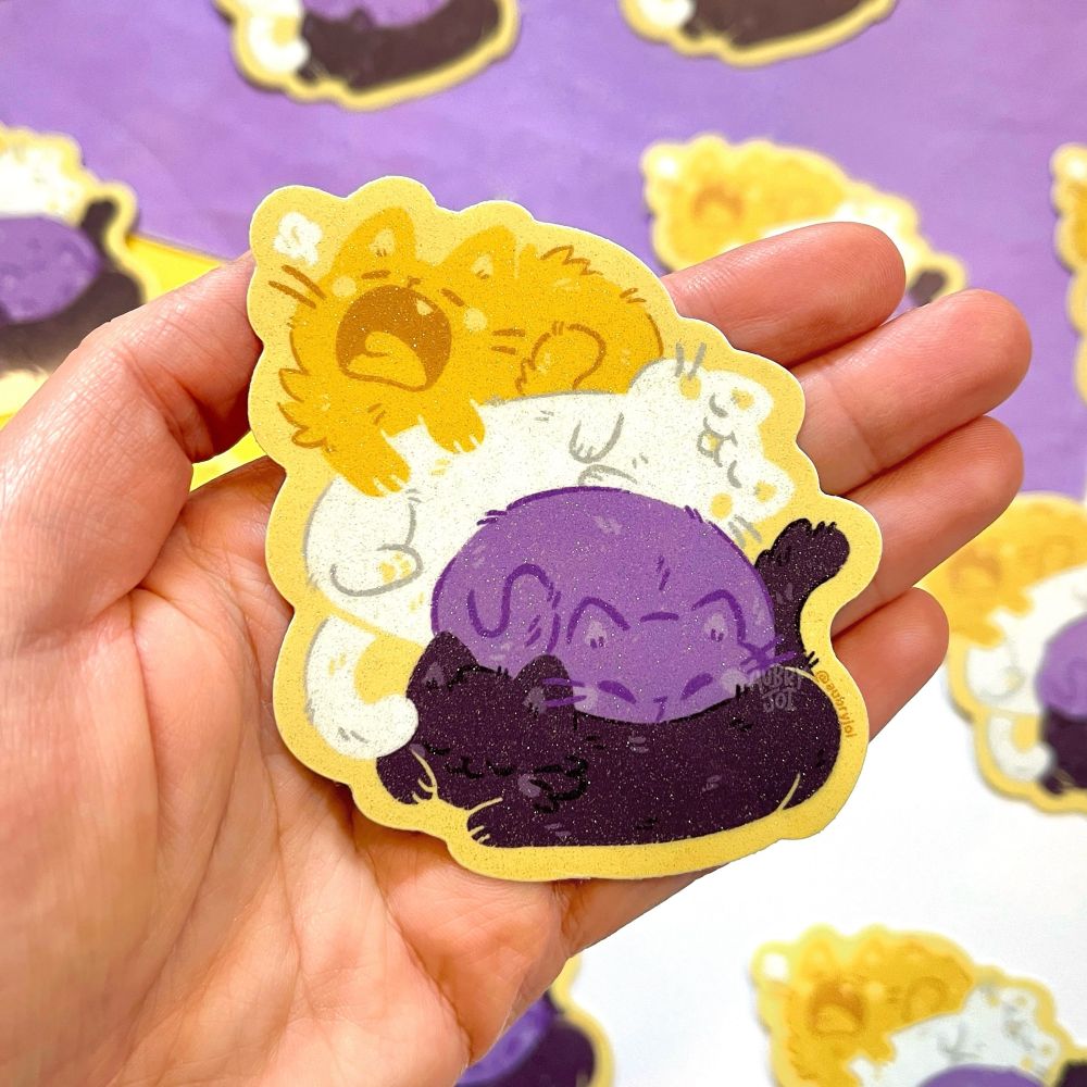 A hand holding a sticker showing four cute, stylized cats stacked and all very sleepy. They are each one color of the non-binary flag, in order: yellow, white, purple, black. The sticker has a glittery coating.