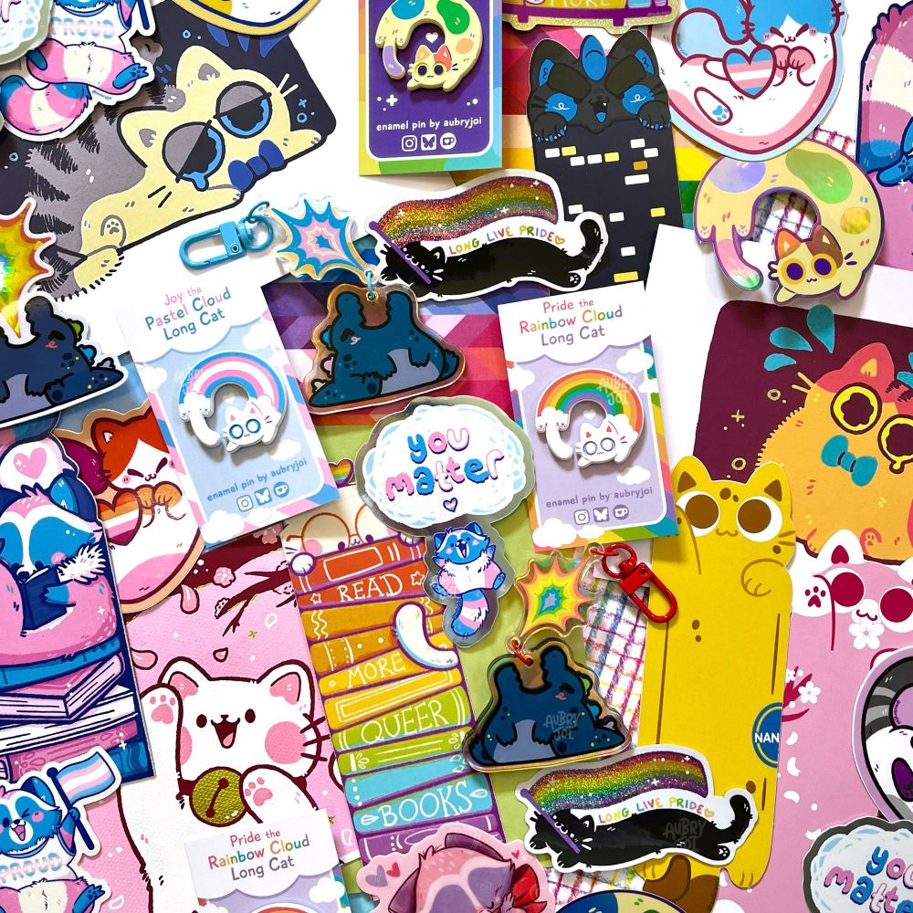 A large spread of assorted merchandise by Aubry Joi, including enamel pins, bookmarks, stickers, art prints, & keychains. Most items feature a cat or Egg the Trans Raccoon. Some feature a chibi Godzilla. Almost everything is themed after pride or pride flags in some way, and it’s all very colorful & cute.
