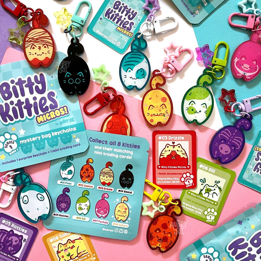 A spread of little cat keychains and mystery bags from the Bitty Kitties Micros collection. 8 different cat charms can be seen, and all are chubby and hanging by their tails. Each has a unique design. The bags are teal and branded for the collection. Each keychain has a colorful clasp and star charm attached to it. There are also tiny trading cards featuring the characters in the spread.