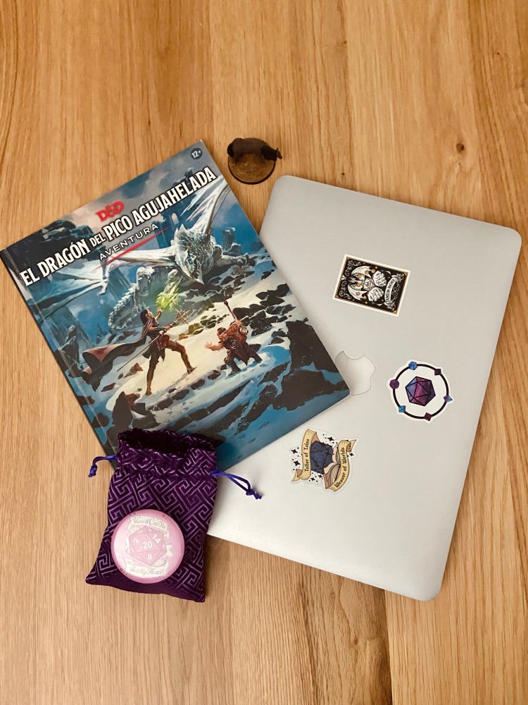 Una mesa sobre la cual hay un libro de la campaña de D&D "El Dragón del Pico Agujahelada", un portátil con pegatinas de rol, un saquito morado de dados, y una miniatura de un caballo marrón.