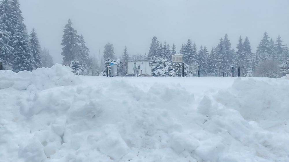 Winterlandschaft mit tiefem Schnee, umgeben von verschneiten Tannenbäumen und der Wetterstation Neuhaus im Hintergrund, Himmel bewölkt.