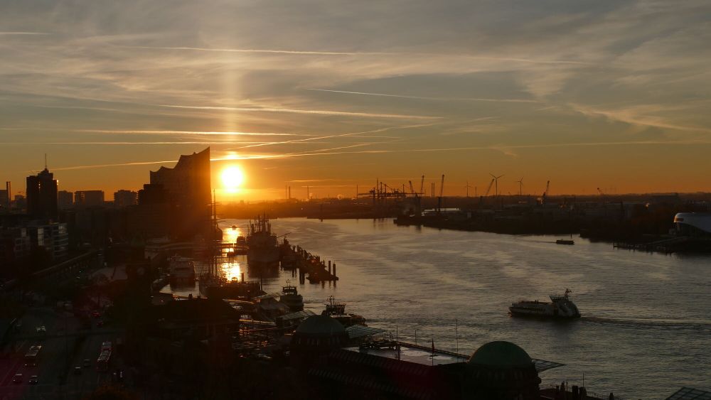 Sonnenuntergang über dem Hamburger Hafen, mit der Silhouette der Elbphilharmonie und Kräne im Hintergrund. Ein Schiff fährt auf der Elbe.