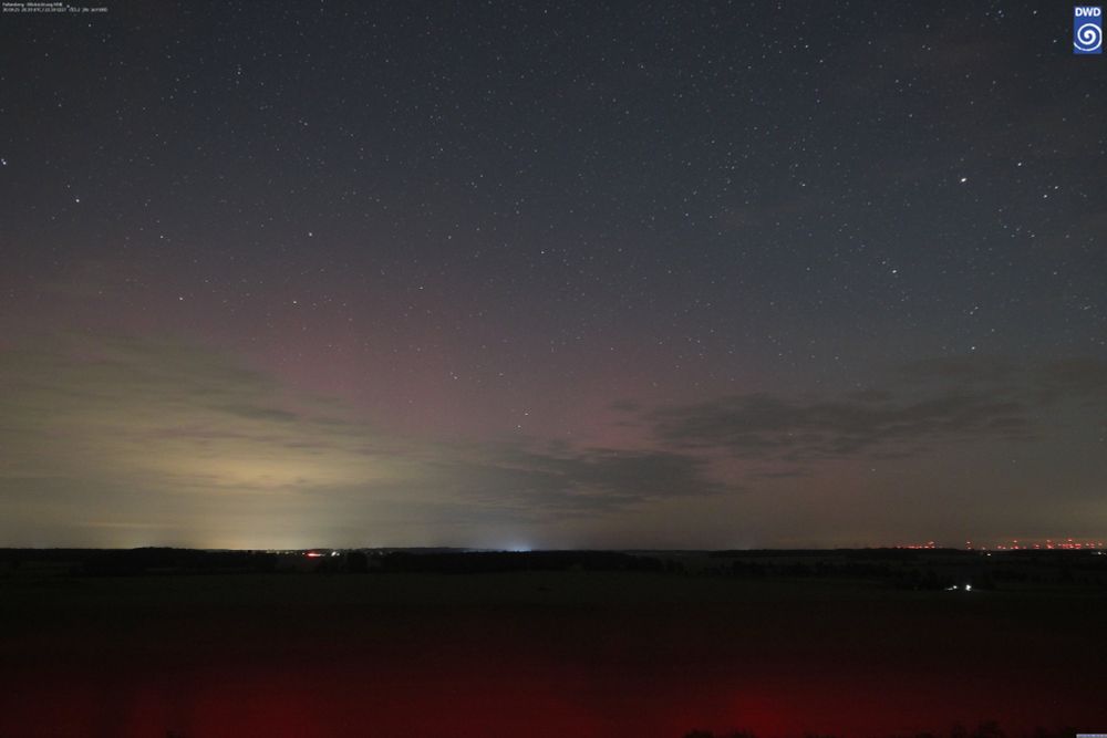Nachthimmel mit Sternen und schwachem Polarlicht über einer dunklen Landschaft, beleuchtet von entfernten Lichtern.