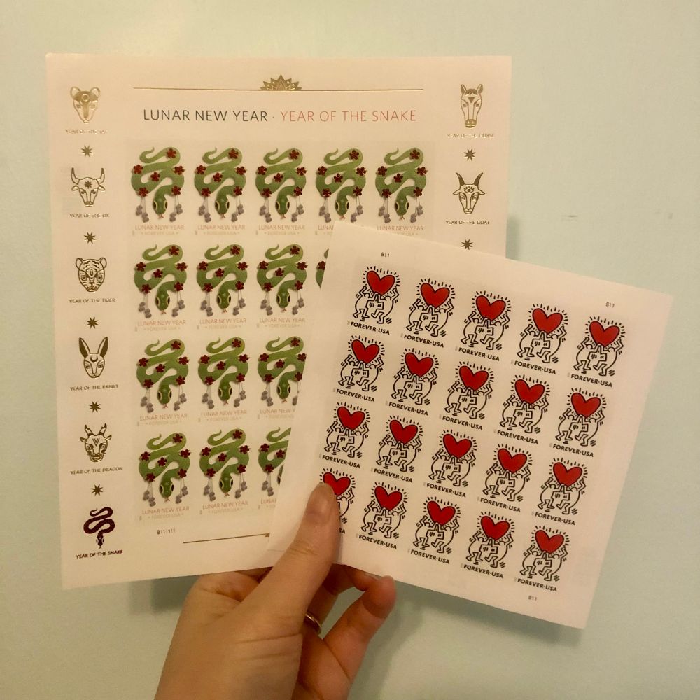Hand holding two sheets of United States Postal Service stamps. Left stamp is Lunar New Year - Year of the Snake stamps, right is Keith Haring Love Stamps