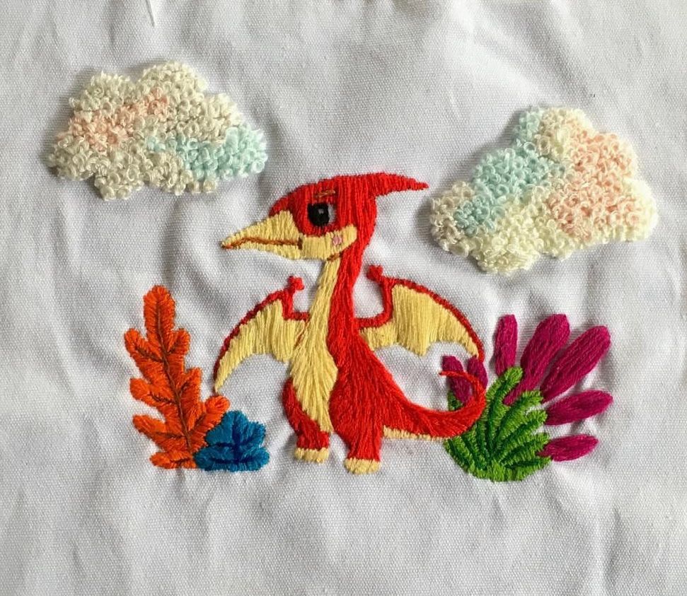 #handembroidery of a red pterodactyl with a yellow tummy. He is standing under cotton candy blue, pink, and cream clouds. And he is surrounded by various colors of plants 