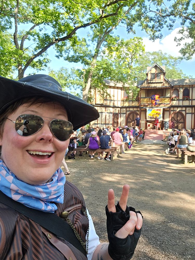 A selfie of me in front of a stage show at the Renaissance Faire. I'm giving a peace sign while wearing a black cowboy hat, a blue and pink bandana, a black and brown striped vest, a white shirt, aviators,  and black fingerless gloves. I'm smiling widely, even though my eyes are hidden by the mirrored lenses on my sunglasses