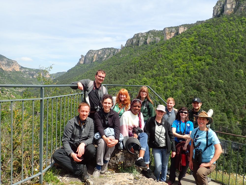 A group of staff and students stood smiling at the camera. There is a gorge in the background with slopes covered in trees and emergent limestone cliffs above.