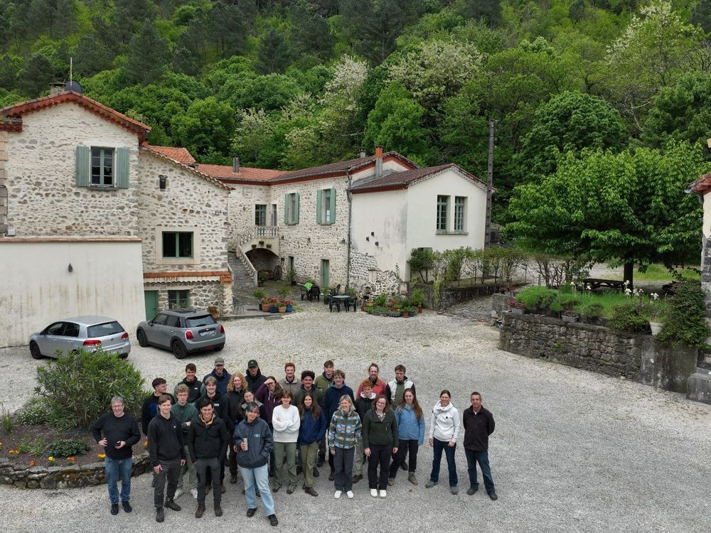 A group of people in front of a traditional building. In the background there is woodland with a mix of tree species in it.