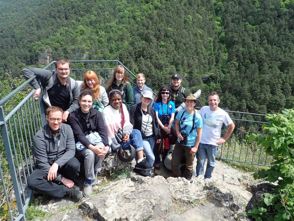 A group of people smiling at the camera. They are stood on a scenic viewpoint with a wooded valley in the background.