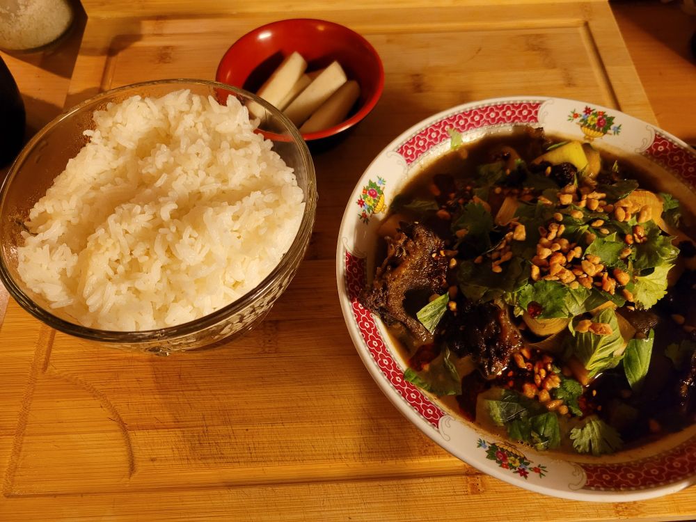 A plate containing a beef and pork stew with herb and chili oil and fried garlic on top, with a side of rice and pickled radish