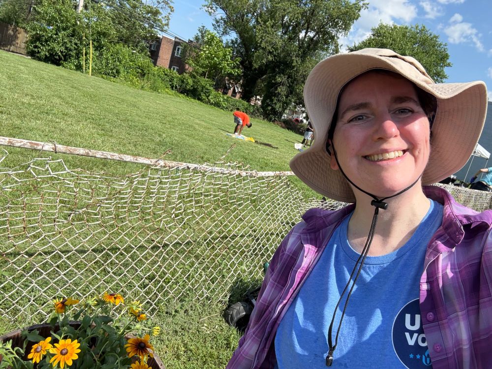 A white person with short dark hair wearing gardening clothes and a sun hat poses with newly transplanted flowers 