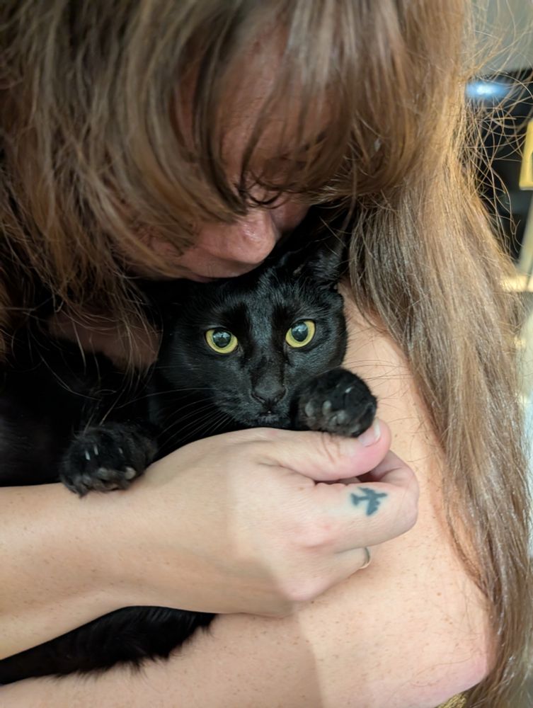 A brown haired femme cuddles a black cat whose expression is both annoyed and pleased.