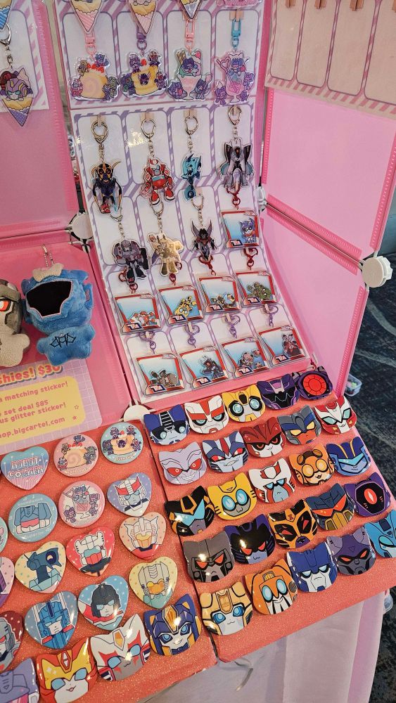 A photo of a portion of an Artist’s Alley booth setup, focusing on acrylic charms and buttons shaped like cat faces. The merch is all of Transformers characters, particularly Transformers Animated.