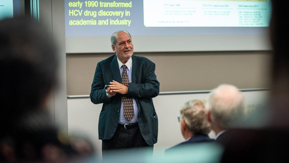 Professor Raymond Schinazi giving a presentation about HCV drug discovery in front of an audience in a conference room. The slide behind shows a timeline and text related to the topic.