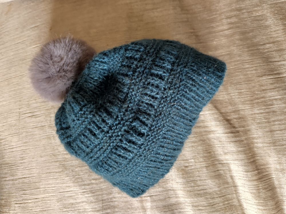 Teal coloured hand knitted hat in alpaca yarn with a mousey grey fake fur pompom. It's laid on a light grey velvet cushion. 