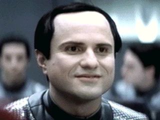 Mathesar from Galaxy quest. He appears to be a middle-aged man with uniformly pallid skin and jet black hair, with a receding hairline. He has a vague, befuddled expression on his face, with a slight smile.