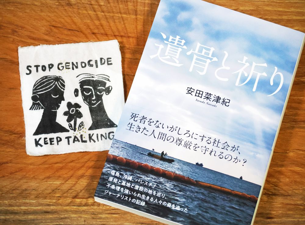 ALT　
『遺骨と祈り』表紙
文字情報：遺骨と祈り　安田菜津紀　Yasuda Natsuki 死者をないがしろにする社会が、生きた人間の尊厳を守れるのか？福島、沖縄、パレスチナ　原発と基地と虐殺の地を巡り、不条理を強いられ生きる人々の姿を追ったジャーナリストの記録　産業編集センター
写真：辺野古の海に、雲間から光が漏れ注いでいる。フロートの向こうで、巡視船がこちらと並走している。

その横に、「STOP GENOCIDE」「KEEP TALKING」の文字と、花を挟んだ二人の人物が描かれたパッチが置かれている。