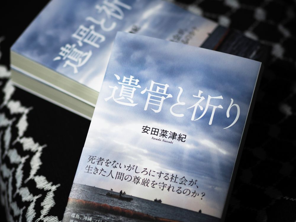 『遺骨と祈り』表紙
文字情報：遺骨と祈り　安田菜津紀　Yasuda Natsuki 死者をないがしろにする社会が、生きた人間の尊厳を守れるのか？福島、沖縄、パレスチナ　原発と基地と虐殺の地を巡り、不条理を強いられ生きる人々の姿を追ったジャーナリストの記録　産業編集センター
写真：辺野古の海に、雲間から光が漏れ注いでいる。フロートの向こうで、巡視船がこちらと並走している。

パレスチナのカフィーヤの上に、手前に一冊、奥に二冊、『遺骨と祈り』が置かれている