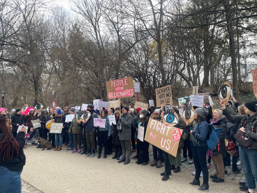 A crowd of 100 people standing together holding signs expressing their dismay with Sen. Schumer 