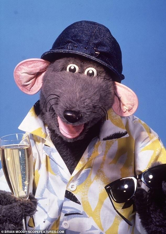 A picture of Roland Rat with sunglasses in one hand and a champagne glass (filled) in the other. 