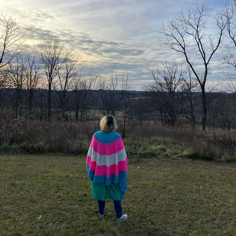 A long haired transmasculine person stands with their bank to the camera, over looking a trap landscape. They are blonde with dark roots, wearing blue jeans and a large handmade crocheted shawl in the colors of the transgender pride flag. The sun is about to set and the trees are barren as it is winter.