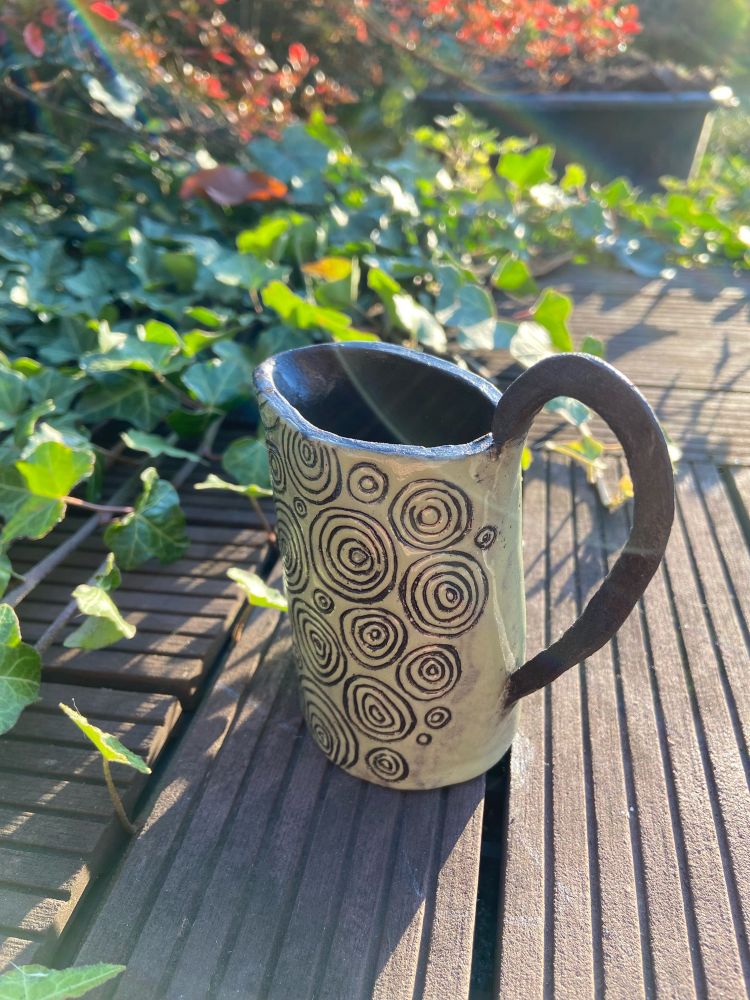 Milchkännchen auf dem Terrassenboden, im Hintergrund ist Efeu. 

Das Kännchen ist aus schwarzem Ton, glasiert in lindgrün mit sgraffito Kreisen. Es hat einen dünnen schwarzen Henkel. 