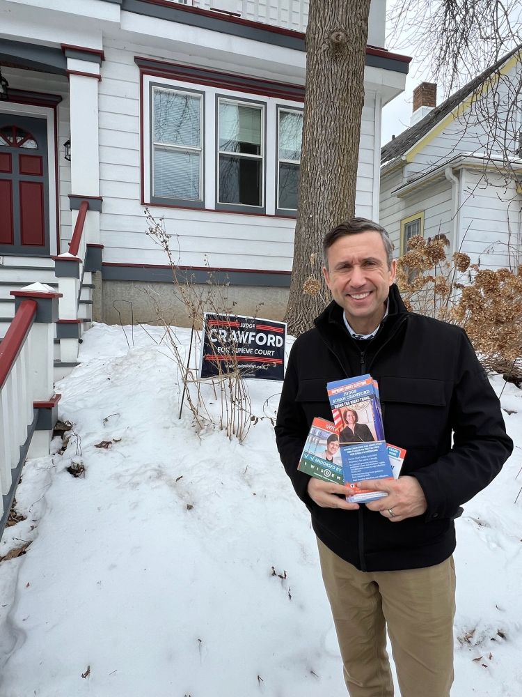 DNC Chair Ken Martin standing in front of a home in Wisconsin, holding canvassing materials for Judge Susan Crawford