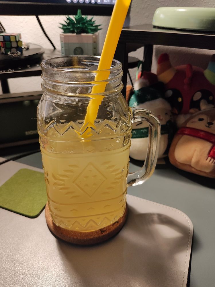 Big glass cup ⅔ filled with water and juice with a yellow silicone straw. Cork coaster underneath it and a felt coaster next to it. Lots of stuff in the background like two wintery small squishmallows an "emotional support demon", green portable speaker, succulent out of lego like building bricks, tiny Rubik's Cube...