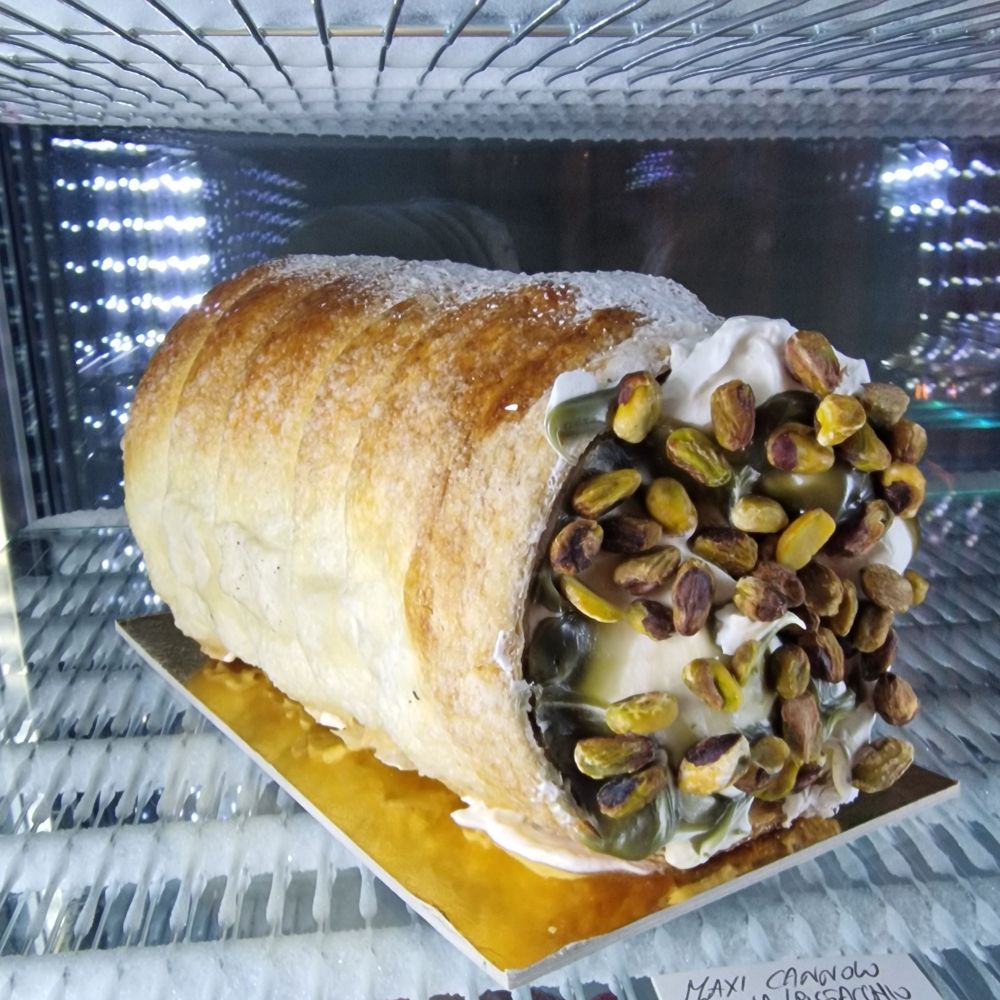 A huge Cannolo, an Italian pastry dessert, widely known after the Godfather movie quote. Has almost 30 cm wide and 50 cm long. 
Made of pastry, icing sugar, cream and pistachios.
Placed inside a fridge with a glass door.