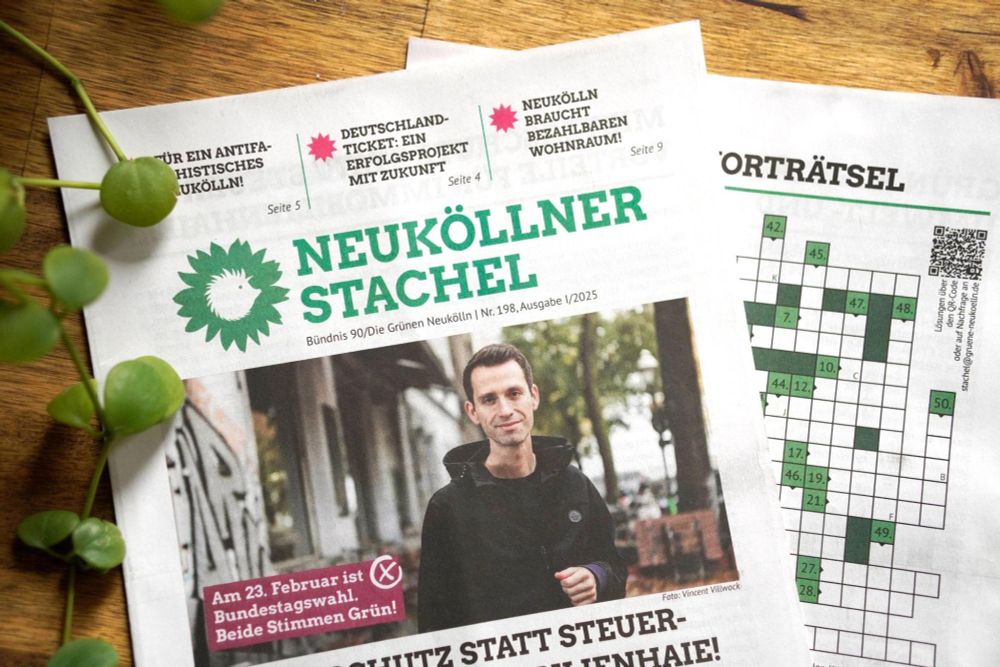 Zwei Exemplare der Bezirkszeitung "Neuköllner Stachel" liegen auf einem Tisch, einmal ist die Vorderseite zu sehen, einmal die Rückseite. Auf der Vorderseite als Titelbild ein Foto von Andreas Audretsch, darin der Schriftzug: "Am 23. Februar ist Bundestagswahl. Beide Stimmen Grün!". Die Rückseite ist größtenteils verdeckt, es ist jedoch ein Kreuzworträtsel zu erkennen.