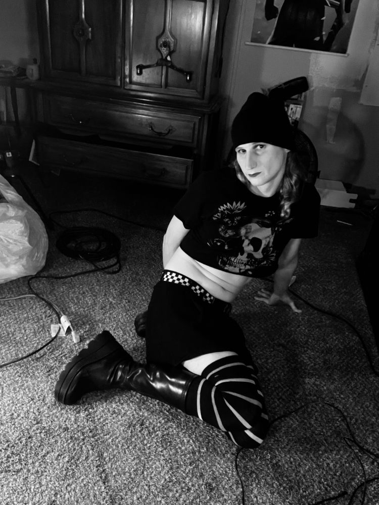 Me sitting on the ground in my hooker boots, striped stockings, black skirt and skater belt with a checkered pattern. My legs are pulled in while my arm holds me up, I'm being sure to bend and show off the curves. On top I'm wearing a black crop top with a skull and flowers design. Lipstick that looks black and a black beanie.