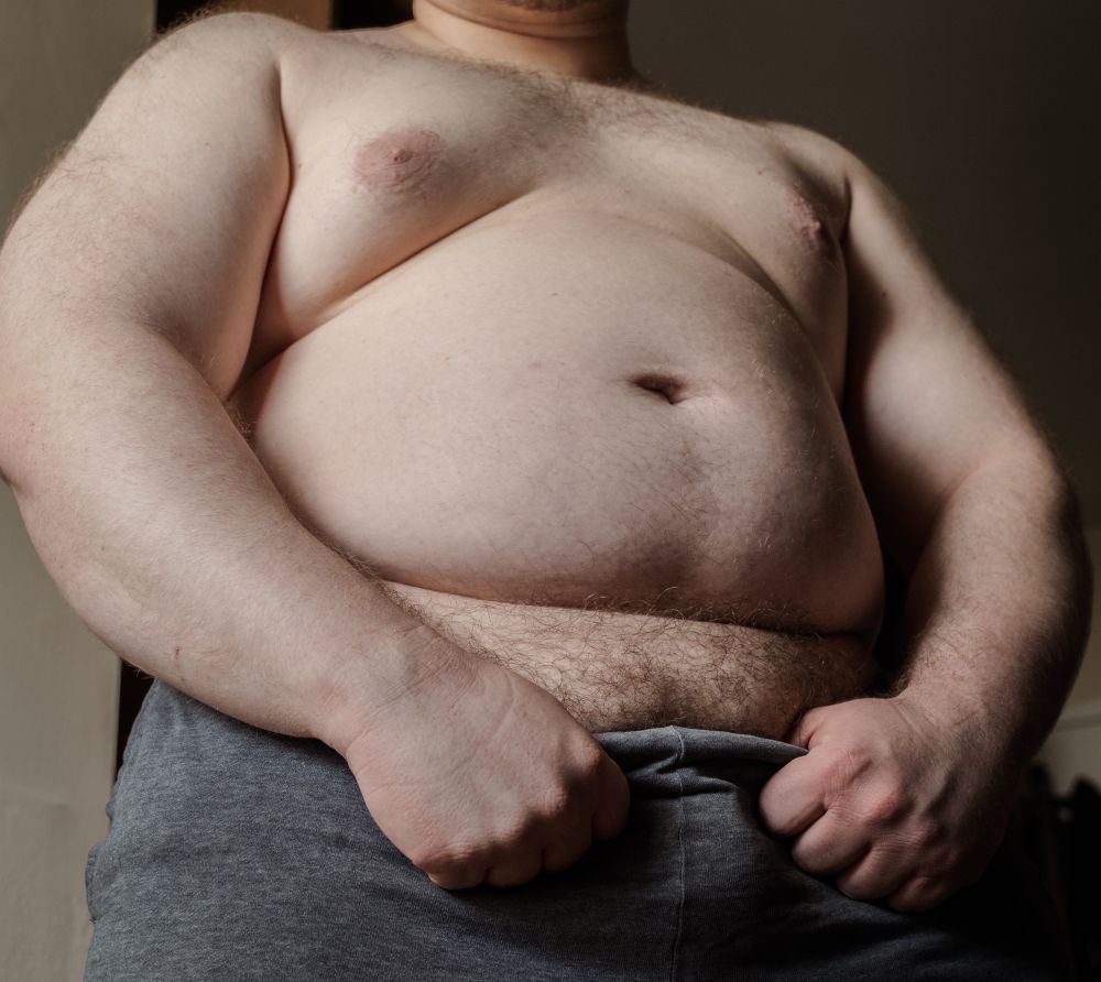A lowered camera angle showing a big male bear presenting his chest and belly to the viewer. His arms outline the sides of his torso, as his hands pull down the waistband of his grey shorts to reveal his pubes.