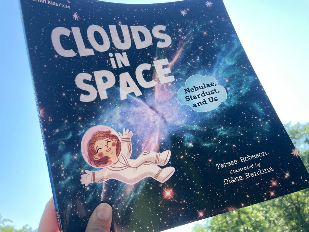 cover of CLOUDS IN SPACE shows a young cartoon astronaut floating in front of stars and a nebula in a black sky; photo is of the book in my hand taken looking up toward a light blue sky with some trees at the bottom