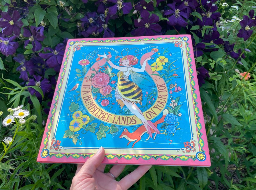 cover of IF A BUMBLEBEE LANDS ON YOUR TOE shows a human-ish bee surrounded by flowers, a fox, and a bird; cover background is blue with pink and yellow edge; book is in my hand; photo background is purple clematis vine, fleabane, and other green leaves