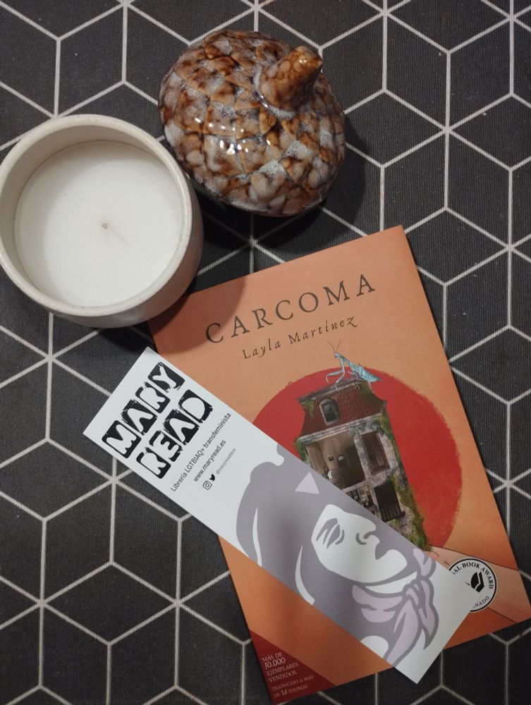 Fotografía del libro Carcoma de Layla Martínez con un marcapáginas de la librería Mary Read y una vela, sobre un hule negro con motivos geométricos blancos