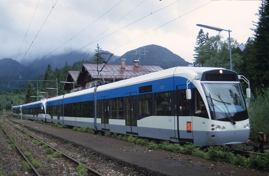 Dreitteilige Saarbahn-Garnitur. Verdeckt ein Gebäude, Berge im Hintergrund, im Vordergrund ein Bahnsteigrest.