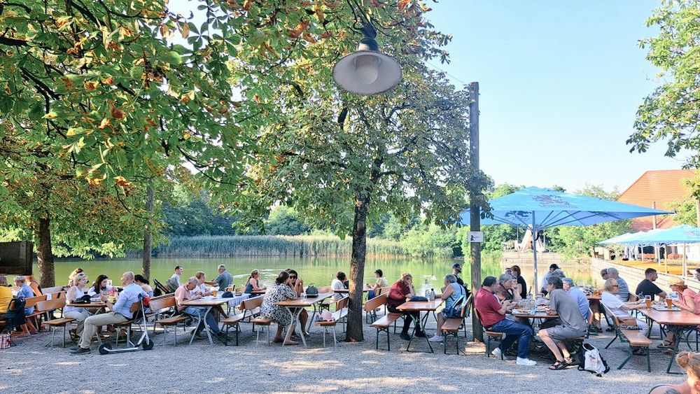 Gut gefüllte Biergartenbänke, Bäume, im Hintergrund See und Parklandschaft.