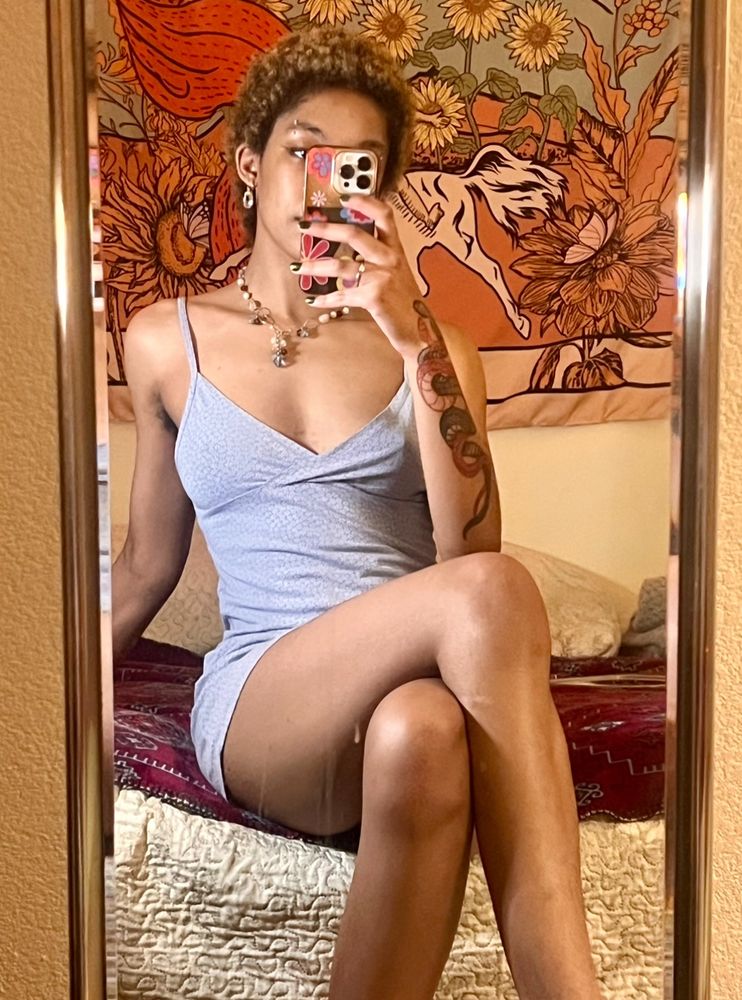 mirror selfie of black girl in a mid length blue dress with a snake tattoo and jewelry and a tarot sun card on the wall