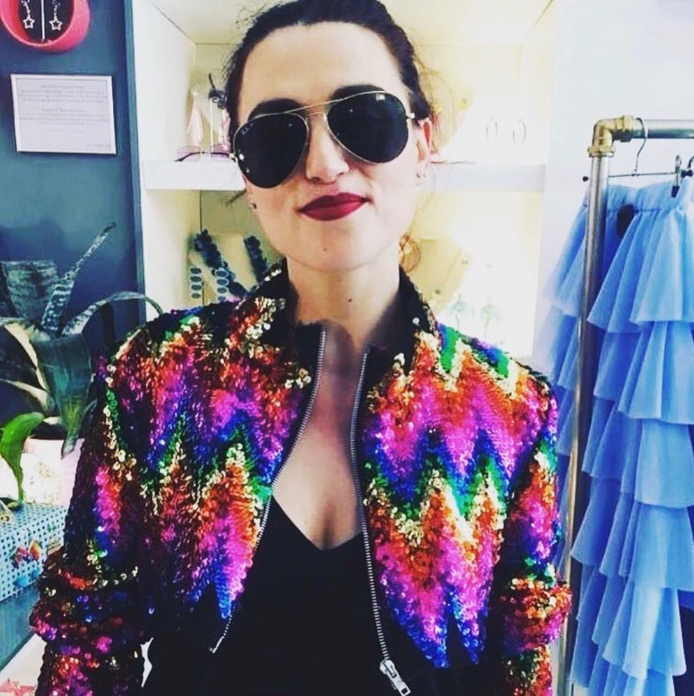 Katie McGrath in sunglasses, a black top and a sequined rainbow jacket. 