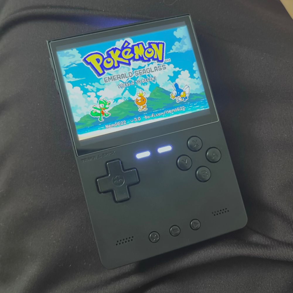 A Trimui Brick handheld device in Black. On the screen is the title screen from Pokemon Emerald Seaglass. 