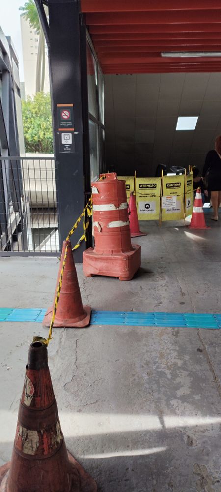 Foto colorida do mostrando que a escada continua parada, lá no fundo, e colocaram mais cones e fitas para sinalizar que o elevador não está funcionando.
