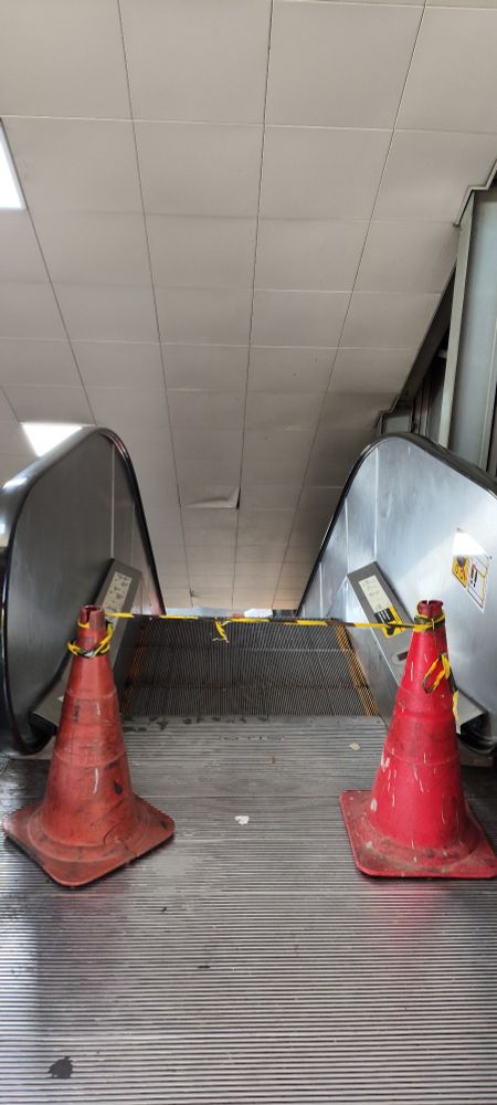 Foto colorida da escada rolante do lado também parada, essa só com dois cones e uma fora preta e amarela impedindo a passagem.