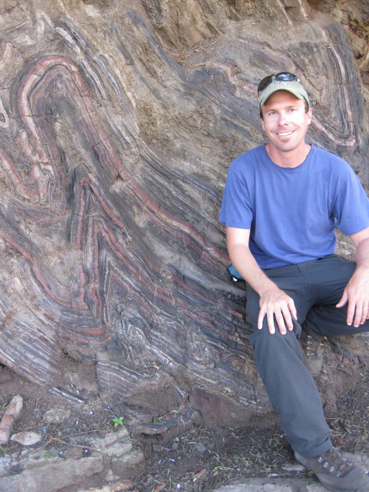 13 years ago, Callan sits on an outcrop of banded iron formation with tight to isoclinal folds of various shapes. He wears a baseball cap with sunglasses perched on top. The rock layers are red, white, and black.
