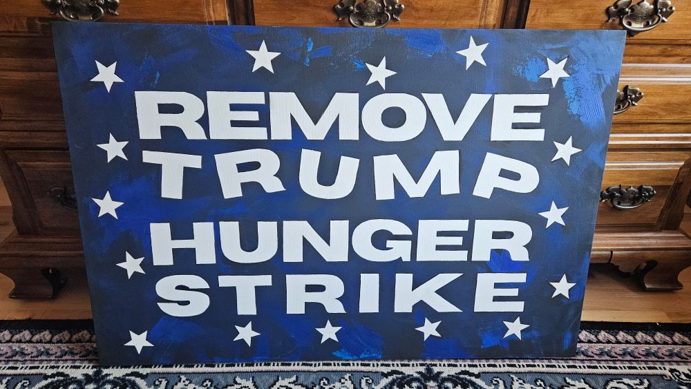 A large 4 foot by 6 foot canvas depicts the words, "Remove Trump Hunger Strike". The background is black and blue and the text stands out in white. It looks unfinished but has the bones of a great composition. Stars circle the words. 