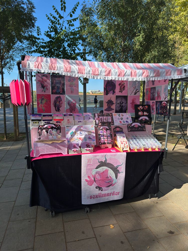 picture shows my artist alley table, in a sunny day outside at Place Saint Pierre, Toulouse France (where I live). The table is covered in a black tablecloth with an illustrated banner, and a bright pink top cloth. I made in black pvc pipes a tabletop four legged structure, that looks like a little vending booth with a striped, pink and white awning at the top. A bright pink decorative paper lantern is hanging on the left of the structure. From there are hanging my big prints, also creating the illusion of curtains. The whole thing is light pink and black, and I think I did a really good job. This took me months to plan, design, build and setup. I made it so that it’s light and compact when transported, but can still look personal and impressive once set up. The entire thing, including displays and stocks, can fit in just two suitcases, so it’s very transportable as well 😌 