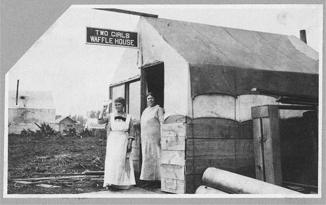 b&w photo of a shack witg the sign TWO GIRLS WAFFLE HOUSE. Two women stand in the doorway wearing dresses with long aprons