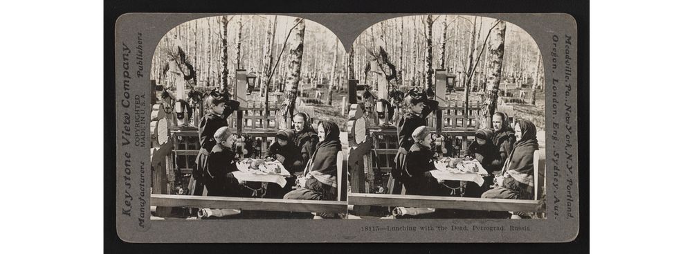Women and children having a meal on a table next to cross-shaped gravestone, in a cemetery.
